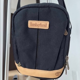 Timberland經典黑色帆布斜背包，搭配棕色皮革飾邊，時尚實用。